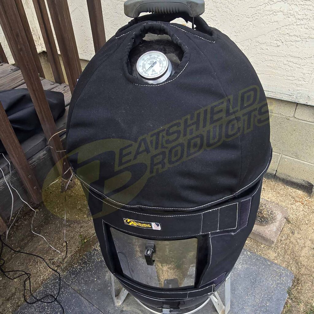 The BBQoozie, a BBQ smoker with a black insulated Heatshield Products jacket and built-in top thermometer, features a heat shield for protection while sitting on a concrete patio near a beige wall.