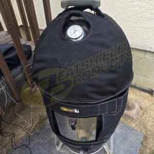 The BBQoozie, a BBQ smoker with a black insulated Heatshield Products jacket and built-in top thermometer, features a heat shield for protection while sitting on a concrete patio near a beige wall.