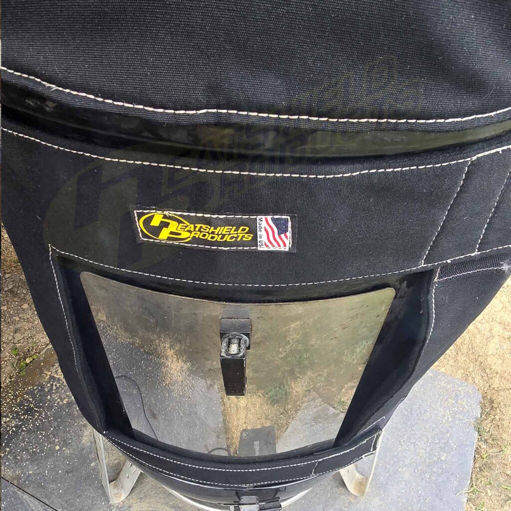 A close-up of the BBQoozie shows a black heat shield cover with white stitching, a yellow Heatshield Products logo, and an American flag patch. Designed for BBQ or smoker setups, it features a clear plastic window and sits on a metal frame.