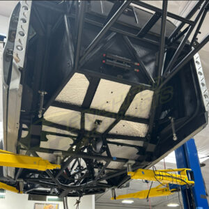A view from below a car lifted on a yellow hydraulic lift, revealing the undercarriage with Sticky Shield® protective panels, including floor pans and transmission tunnels, in a garage or workshop setting.