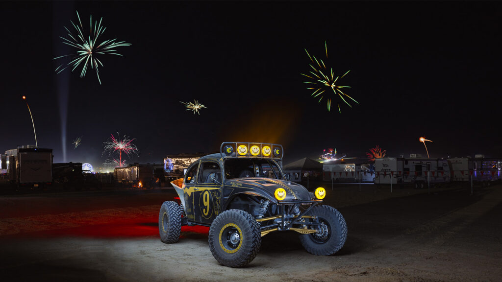 Black and yellow Baja Bug with KC lights under fireworks at a nighttime off-road event