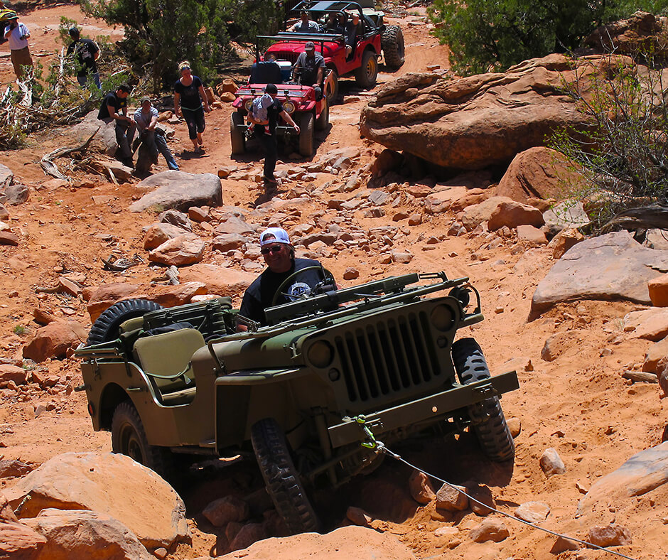 A green Jeep gets towed up rocky path as others off-road behind him