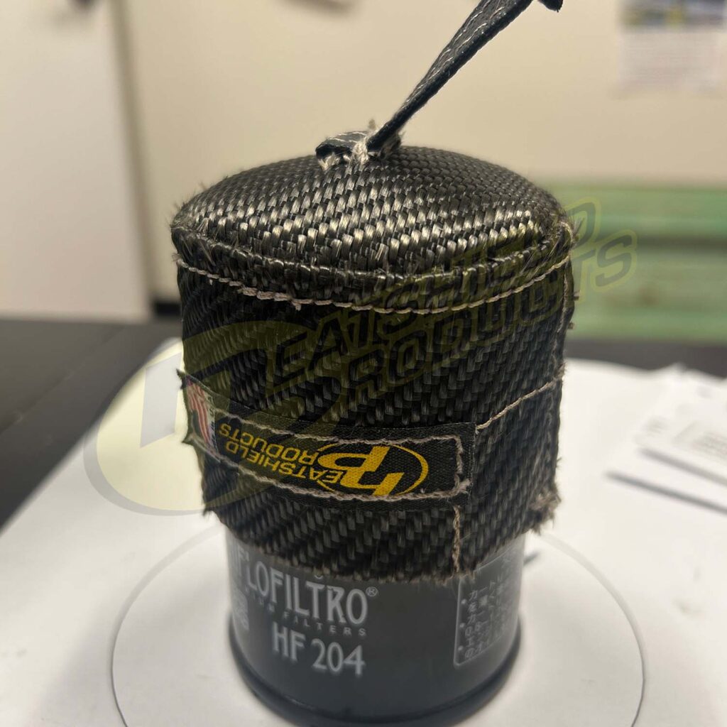 A cylindrical canister labeled Custom Solutions is wrapped in a black, carbon-fiber-like material with a Velcro strap and a tag reading EASTSHIELD PRODUCTS, sitting on a white surface.